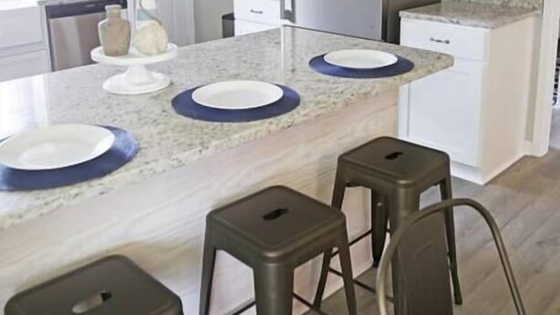 A modern kitchen with white cabinets, a stainless steel refrigerator, a marble countertop, and three bar stools. - Gulf Coast Condos on the Sand Dunes - 202 (Dauphin Island)