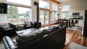 A modern living room with a large sofa, a TV, and a fireplace. There is a kitchen area with a stove and microwave. The room has large windows and a balcony.