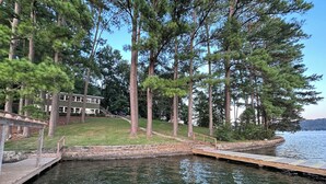 A dock extending into a calm lake, surrounded by tall pine trees and a building with a sloped roof. - Guntersville Waterfront Cabin - Panoramic View - Large Dock (Guntersville)