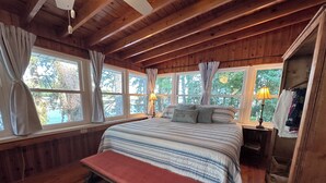 A wooden cabin bedroom with a large bed, a bench, and multiple windows offering a view of trees. - Guntersville Waterfront Cabin - Panoramic View - Large Dock (Guntersville)