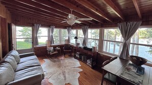 A cozy cabin interior with a wooden ceiling, large windows overlooking a lake, a leather couch, a dining table, and a fireplace. - Guntersville Waterfront Cabin - Panoramic View - Large Dock (Guntersville)