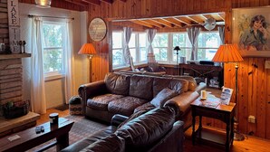A cozy cabin interior with a leather sofa, wooden walls, and a large window offering a view of trees. - Guntersville Waterfront Cabin - Panoramic View - Large Dock (Guntersville)