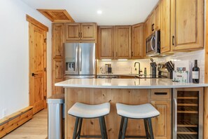 A modern kitchen with wooden cabinets, stainless steel appliances, a large island with stools, and a wine fridge.