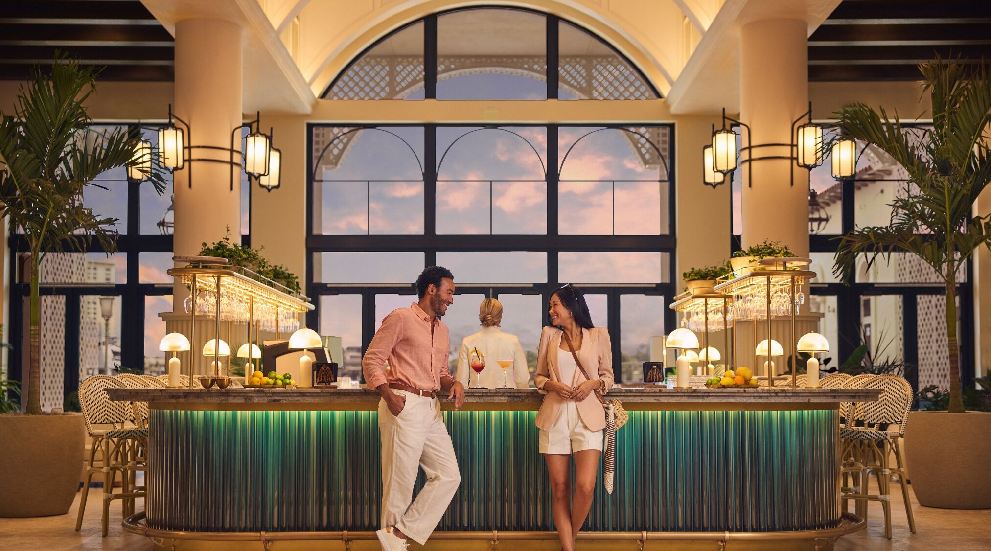 Zona de bar con vistas al cielo a través de amplios ventanales, una barra con fondo iluminado en verde y sillas de mimbre.