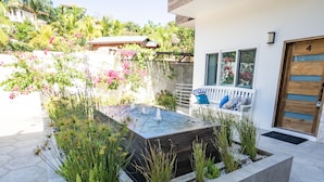 Patio moderno all'aperto con un elemento d'acqua rettangolare, circondato da diverse piante e un'area relax con una panchina e dei cuscini.