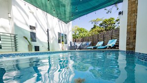 Una piscina con acqua cristallina, circondata da un edificio bianco con ampie finestre e un'area coperta con lettini.
