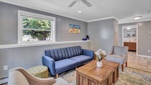 Ein Wohnzimmer mit einem blauen Sofa, einem Holztisch, einem beigen Sessel und einem grauen Sessel. Ein Fenster bietet Blick auf Bäume, und an der Wand hängt ein Gemälde.