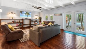 Ein geräumiges Wohnzimmer mit einem großen grauen Sofa, einem Liegesessel und einem Bücherregal. Eine Deckenventilator, ein Fernseher und Blick auf Palmen durch die Glastüren sind vorhanden.