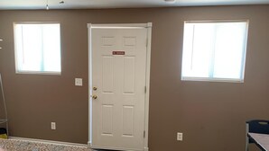 A room with a patterned bedspread, a white door with a 'No Smoking' sign, two windows with blinds, and a small table with a chair. - Desert Oasis Campground (McNeal)
