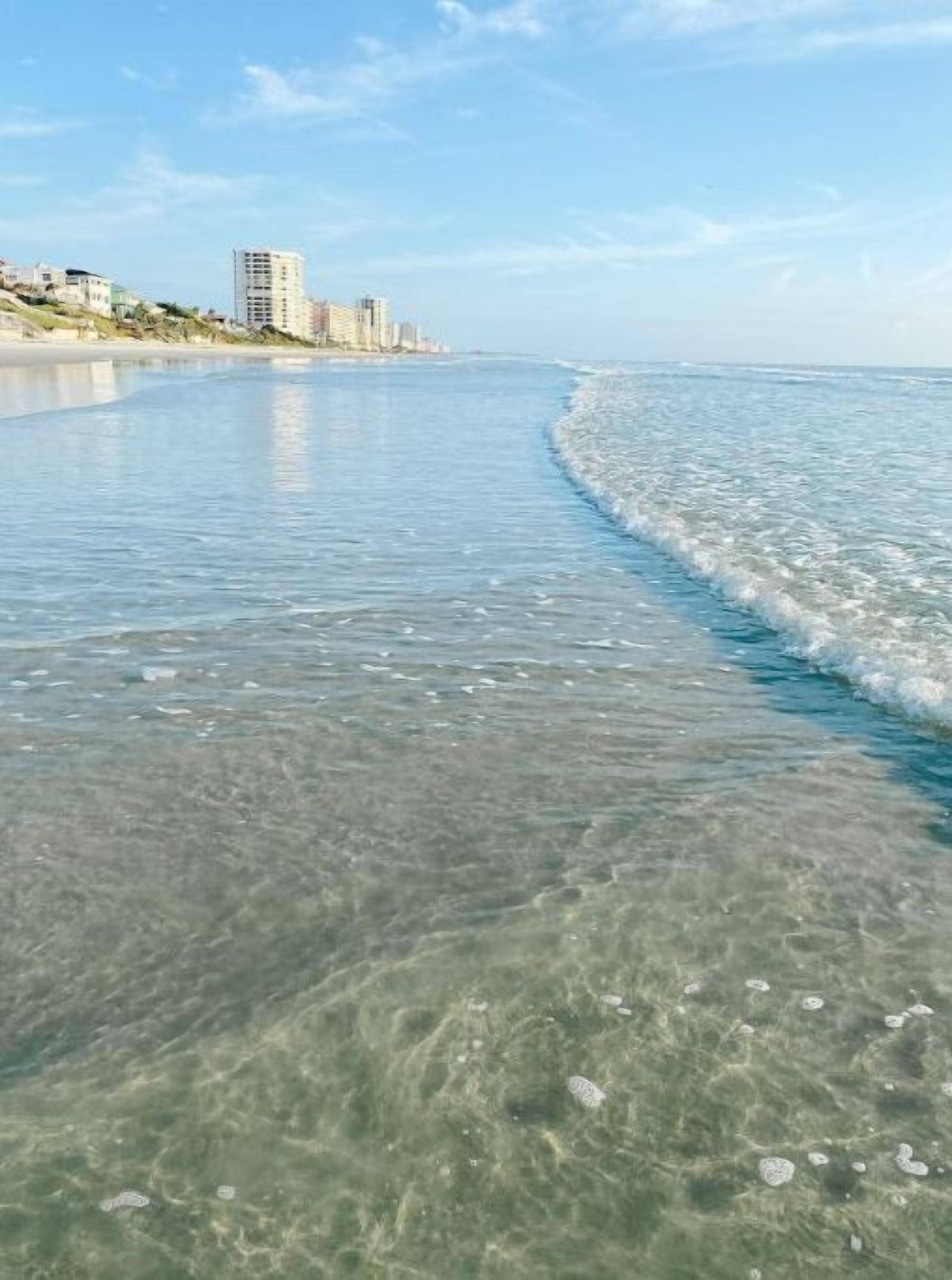 Una playa con aguas transparentes y poco profundas, y una vista distante de edificios en la costa.