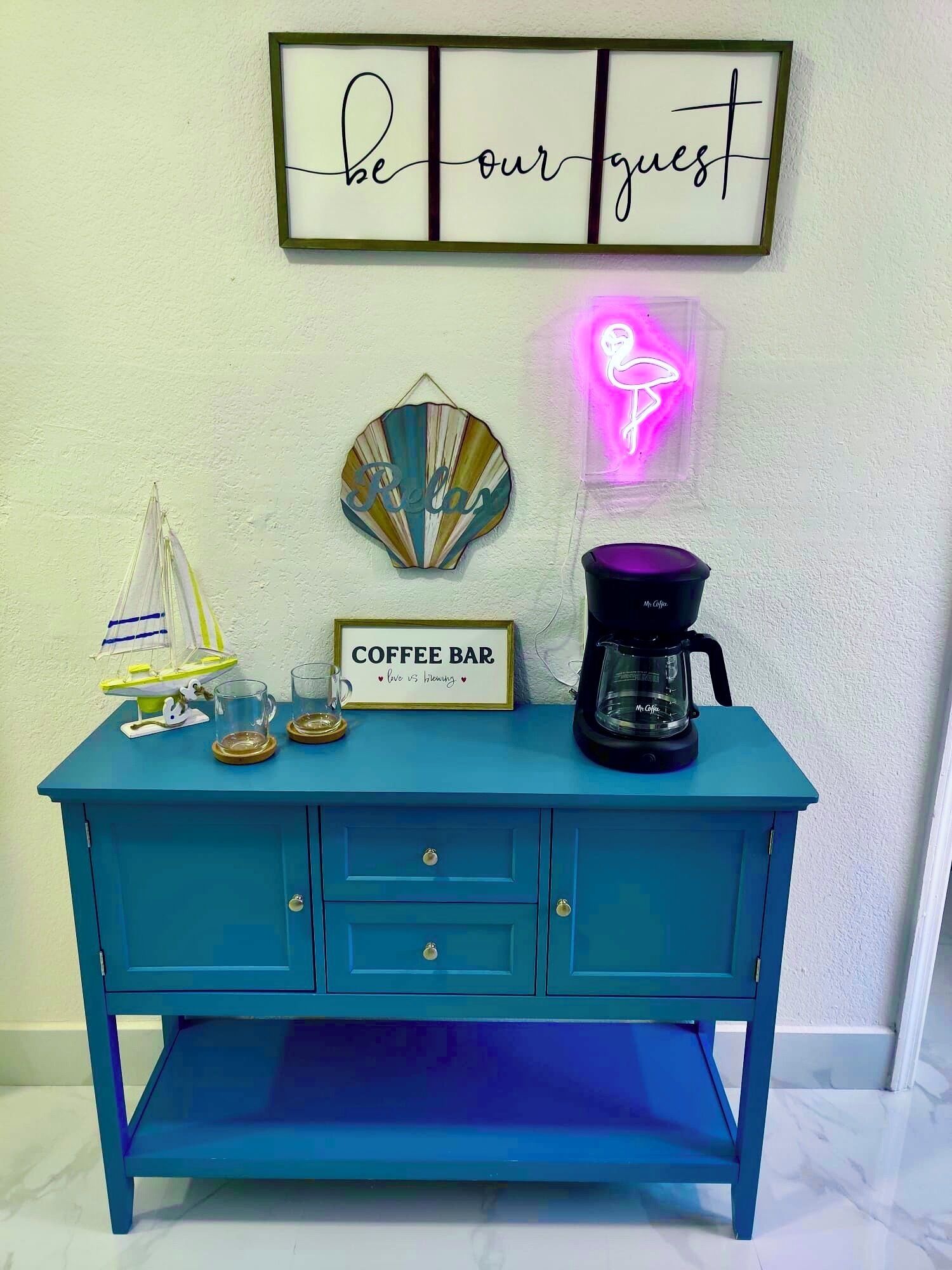 A teal coffee bar with a neon flamingo sign, a sailboat decoration, and a framed 'be our guest' sign.