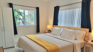 A hotel room with a bed, a nightstand, a lamp, a wardrobe, and a window with curtains. - Beautiful Chic Home Near Coconut Grove & Brickell (Miami)