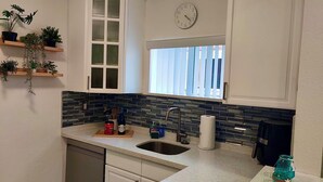 A kitchen with white cabinets, a black tiled backsplash, a window with blinds, a clock, and a sink. - Beautiful Chic Home Near Coconut Grove & Brickell (Miami)