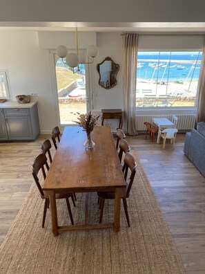 Un coin repas avec une table et des chaises en bois, un vase contenant des fleurs séchées, un miroir au mur et une vue sur la mer par de grandes fenêtres.