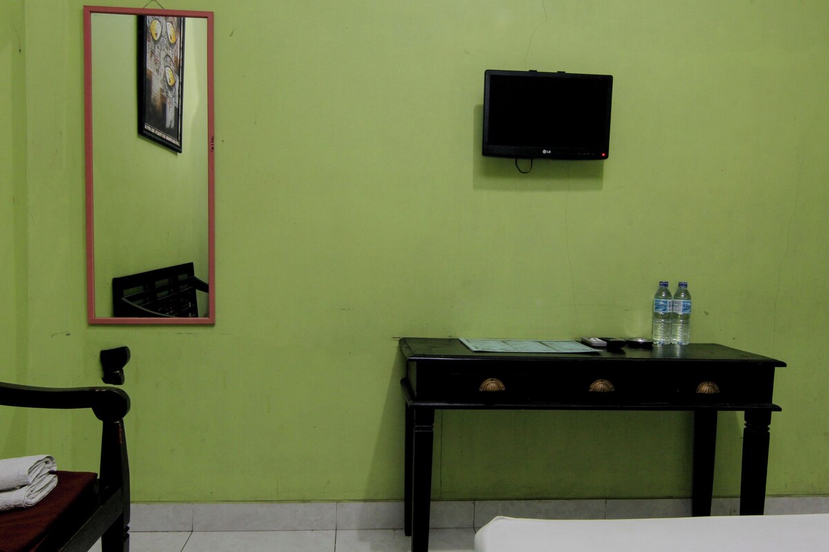 Una habitación con pared verde, un televisor negro montado en la pared, una mesita con dos botellas de agua, una silla con una toalla doblada y una cama con ropa blanca.