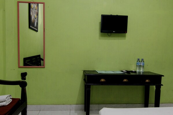 Una habitación con pared verde, un televisor negro montado en la pared, una mesita con dos botellas de agua, una silla con una toalla doblada y una cama con ropa blanca.