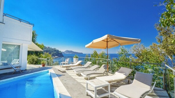 A poolside area with lounge chairs and an umbrella, overlooking a clear blue sea and distant mountains.