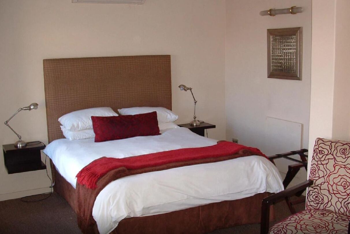 Cama bem arrumada com lençóis brancos e manta vermelha, duas luminárias de cabeceira, uma cadeira estampada e um objeto decorativo na parede.