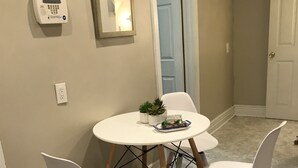 Un petit coin repas avec une table ronde et quatre chaises blanches. Une petite plante en pot est posée sur la table, et une prise de courant est visible sur le mur.