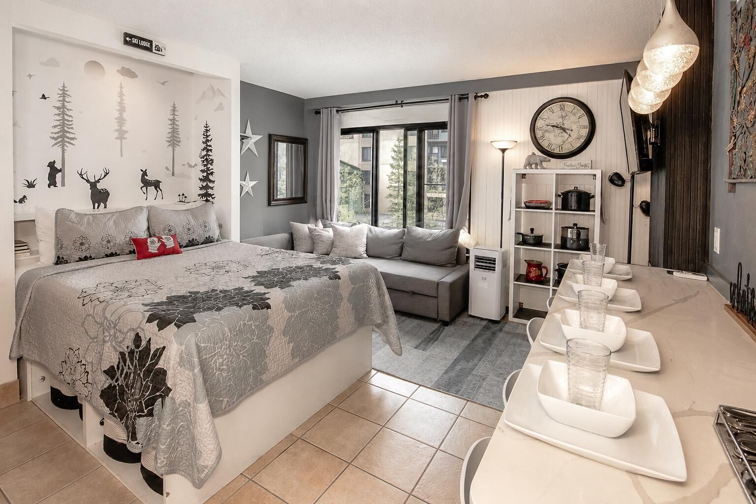 Un dormitorio con cama, sofá, mesa de comedor con vasos, un reloj y ventana con cortinas.