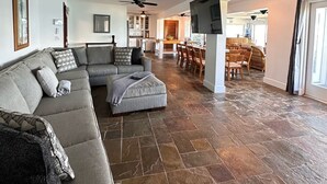 A spacious living area with a grey sectional sofa, a small ottoman, and a ceiling fan. The room has a stone tile floor and a large flat-screen TV mounted on the wall.