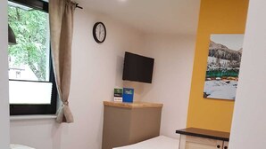 Quarto compacto com cama, uma mesinha, um banco, TV fixada na parede e janela com cortinas.