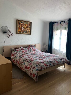 A bedroom with a bed, a wall-mounted fan, a floral painting, and a window with curtains.