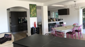 A modern kitchen with dark cabinetry, a dining area with a large table and pink chairs, and a living area with a sofa and a decorative pillow. - Villa 5 Chambres, vue Mer, Piscine, 5´ des Plages et Golf, Terrasses, Parking (Mandelieu-la-Napoule)
