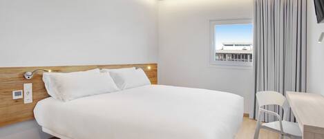 A modern hotel room with a large bed, a wooden headboard, a desk with a chair, and a window with curtains.