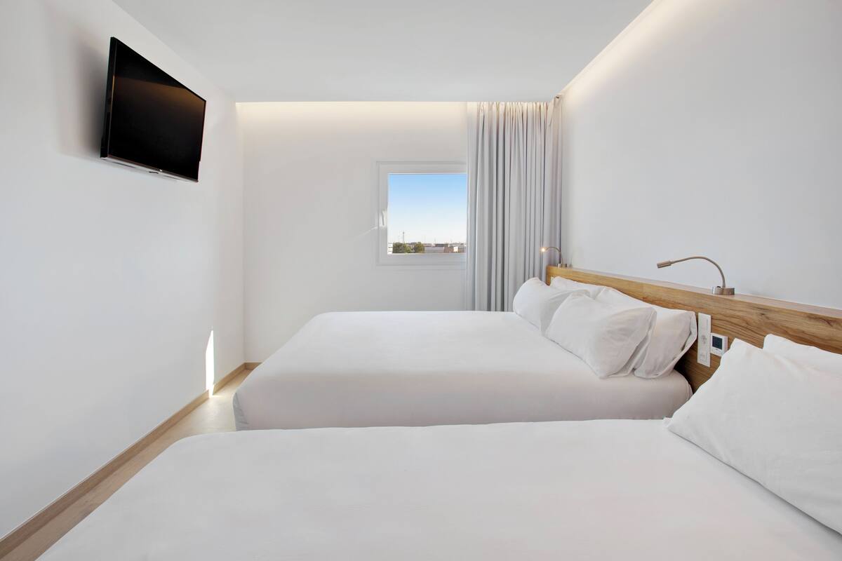 Una habitación de hotel moderna con dos camas, un televisor de pantalla plana montado en la pared, una ventana con cortinas y un cabecero de madera.