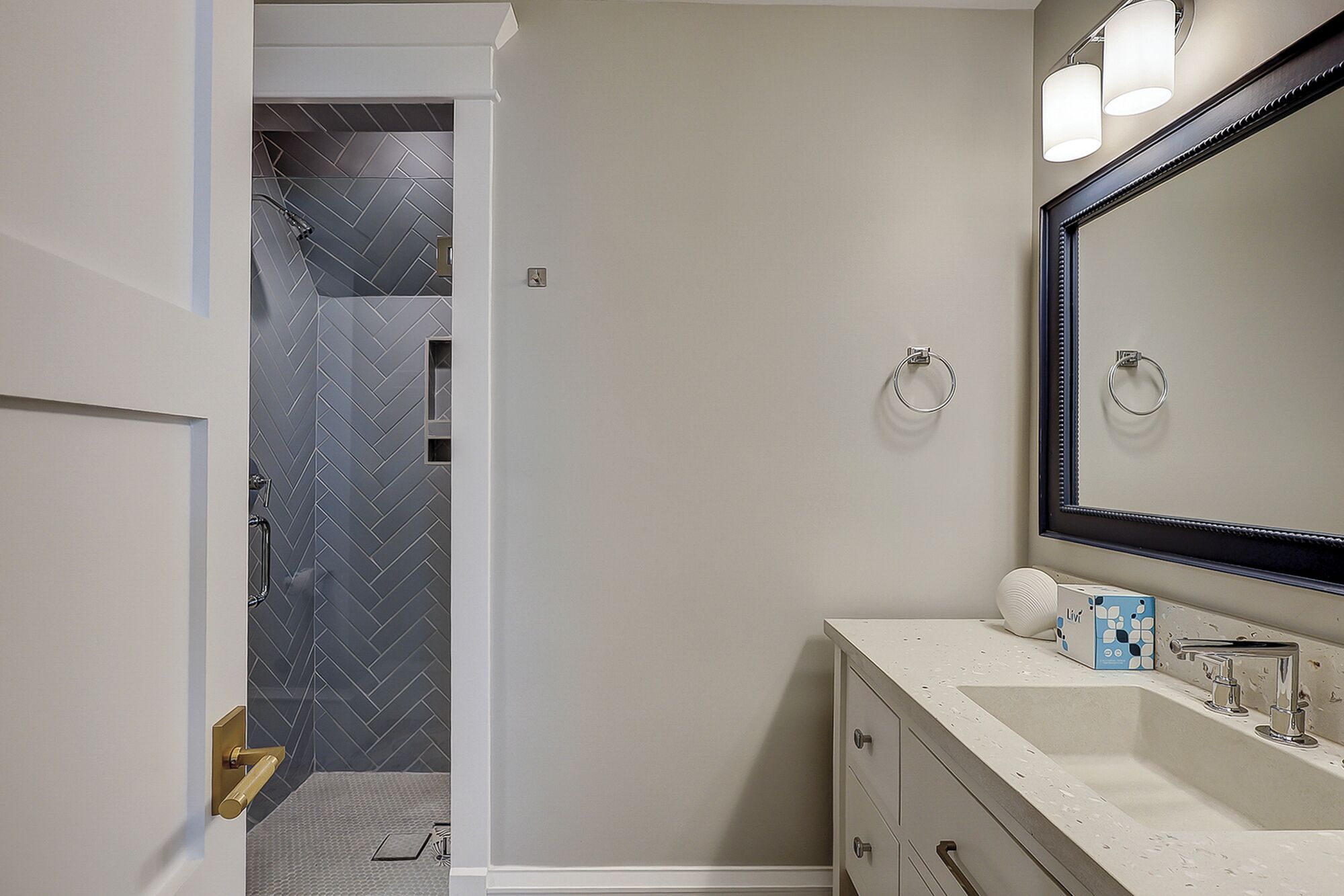 A bathroom with a herringbone-patterned shower wall, a large mirror, and a vanity with a sink and toiletries.