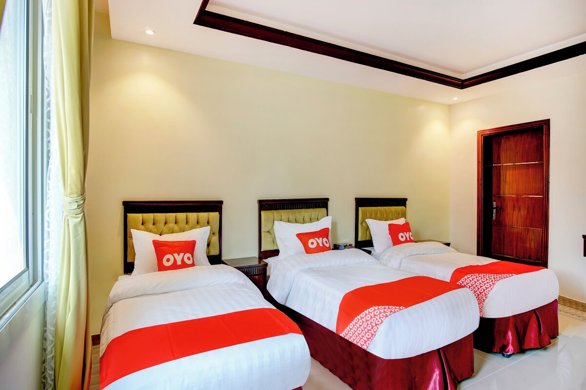 Habitación de hotel con tres camas, cada una con una colcha y almohadas rojas y blancas. La habitación tiene un cabecero de madera, un ventanal con cortinas y una puerta de madera.