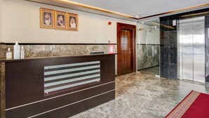 Una recepción con retratos enmarcados, una puerta de madera, un ascensor de vidrio y un piso de mármol.