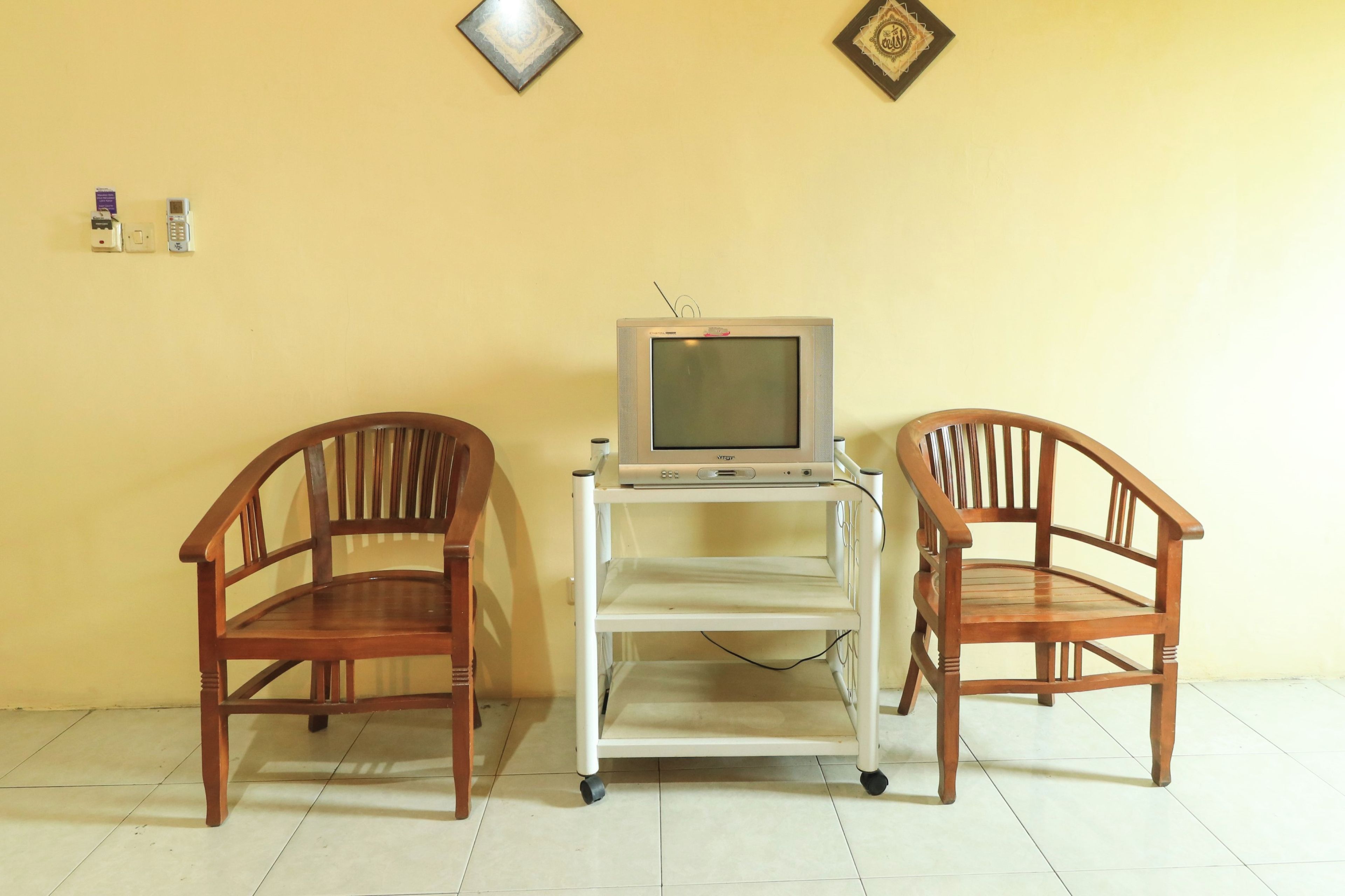 A room with two wooden chairs, a television on a stand, and a wall with two framed items.