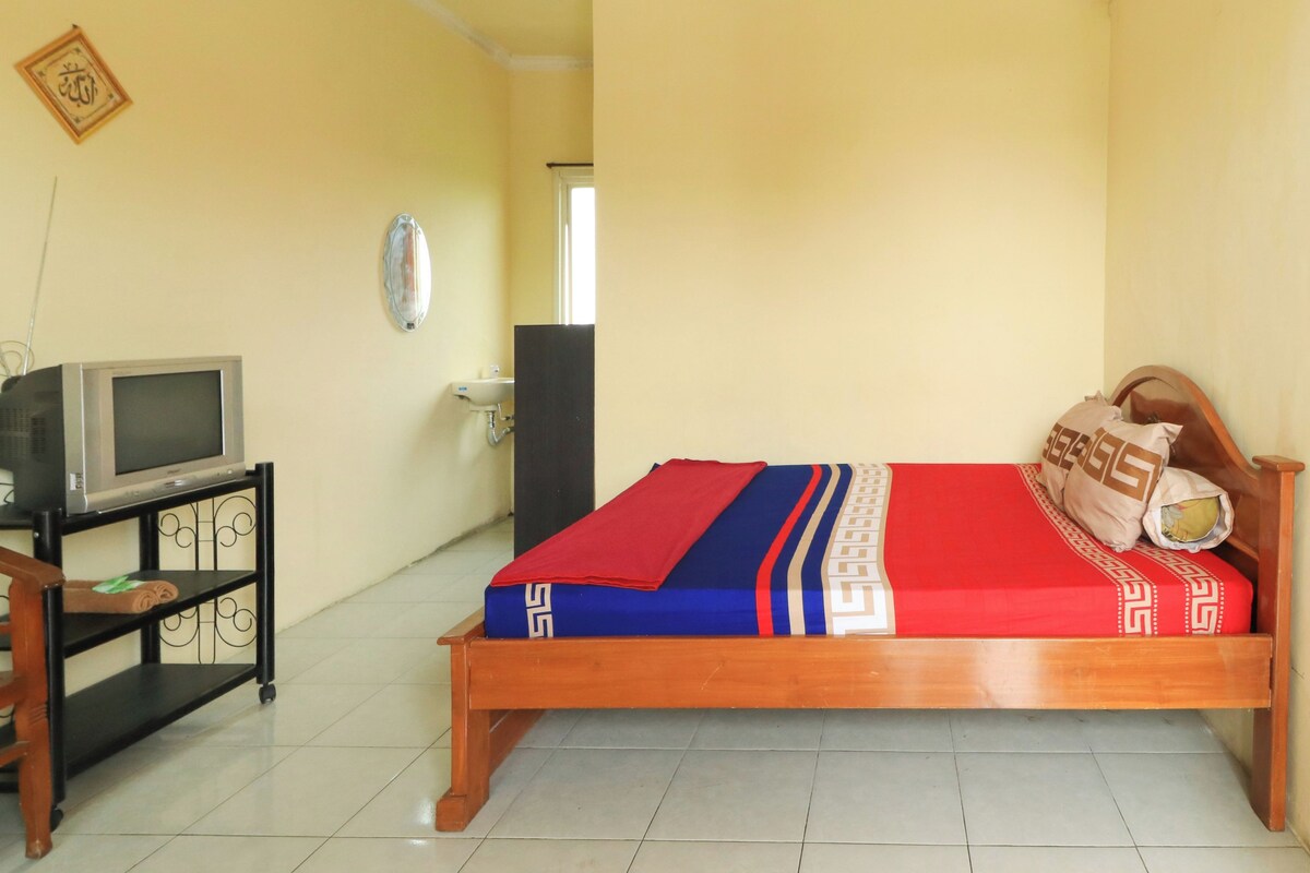 A room with a bed, a television on a stand, a wall clock, and a door leading to another room.