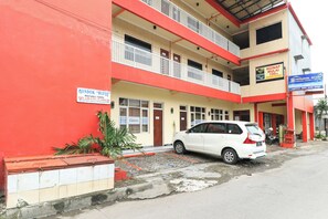 Um edifício de vários andares com fachada vermelha e bege, um carro branco estacionado na frente e um aviso indicando 100% de ocupação.