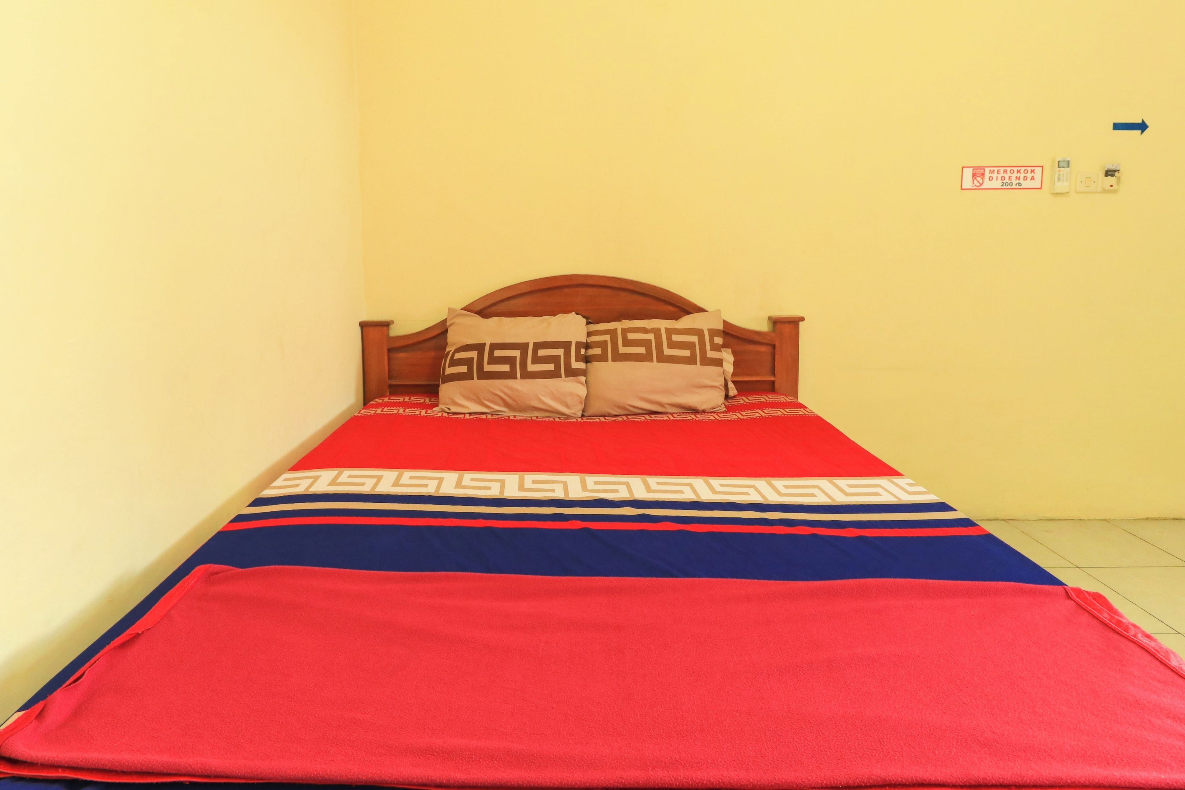 A single bed with a wooden headboard, a red and blue bedspread, and two pillows with a geometric pattern. There is a sign on the wall indicating a no-smoking area.