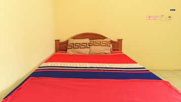 A single bed with a wooden headboard, a red and blue bedspread, and two pillows with a geometric pattern. There is a sign on the wall indicating a no-smoking area.