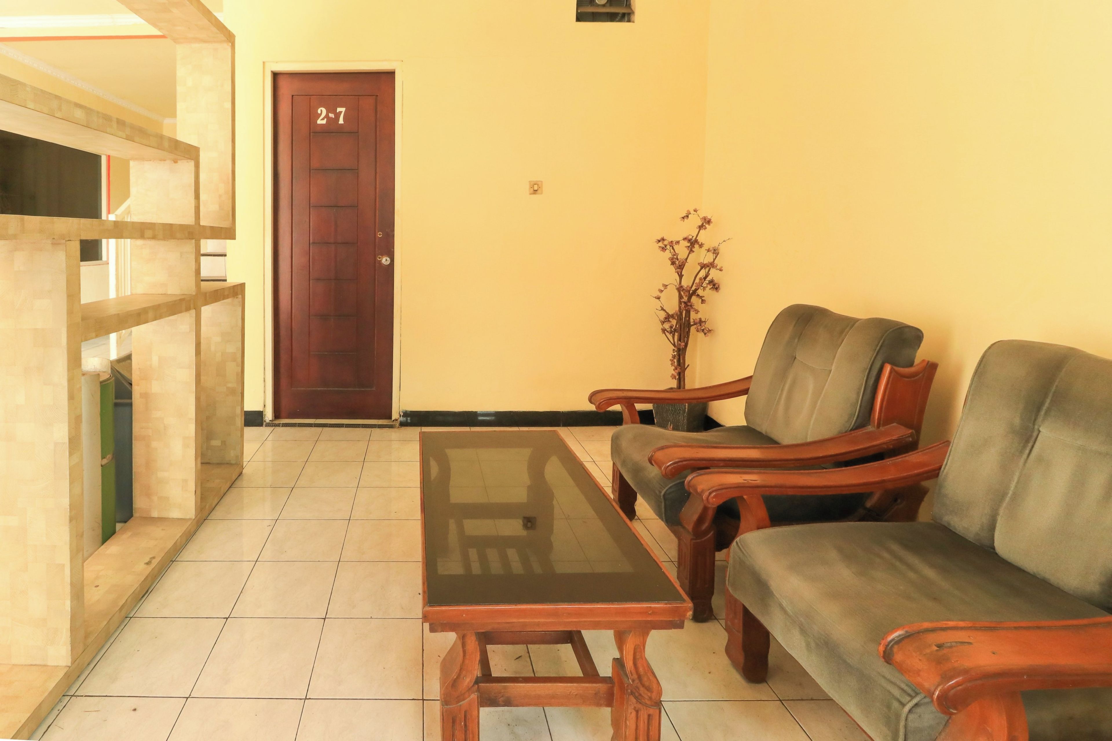 A room with a wooden door marked '2nd 7th', a glass-top table, two wooden armchairs, and a small potted plant.