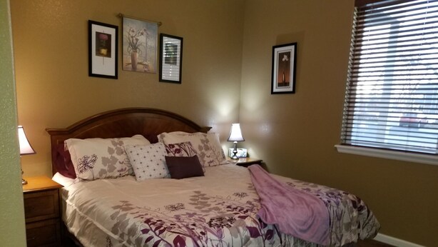 Cama bem arrumada com roupa de cama floral, duas luminárias de cabeceira e quadros nas paredes.