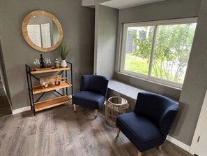 A room with a grey wall, a round mirror, two blue chairs, a wooden shelf with bottles, and a window with a view of greenery.