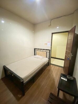 Una habitación pequeña con cama individual, piso de madera, mesita de noche y puerta que da a otra habitación.
