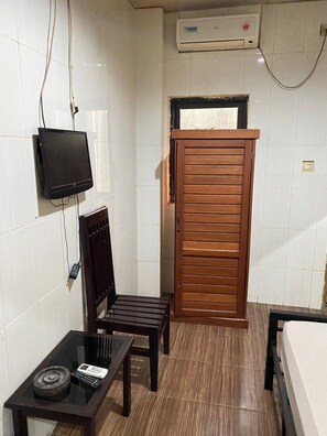 Una habitación pequeña con televisión montada en la pared, una puerta de madera, una mesita con un control remoto y una cama.