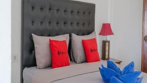 Ein Bett mit einem gepolsterten Kopfteil, zwei rote Kissen, einer beigen Tagesdecke und einem blauen Zierkissen.