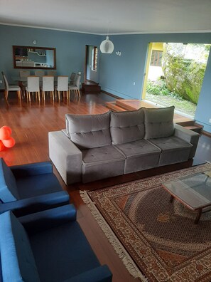 A modern living room with a grey sectional sofa, a blue armchair, and a glass coffee table. There is a dining area with a white table and chairs, and a staircase leading to an outdoor area with greenery.