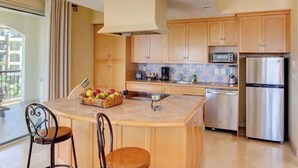 Een moderne keuken met houten kastjes, een roestvrijstalen koelkast, een magnetron en een koffiezetapparaat. Er is een klein eiland met een fruitschaal en twee stoelen.