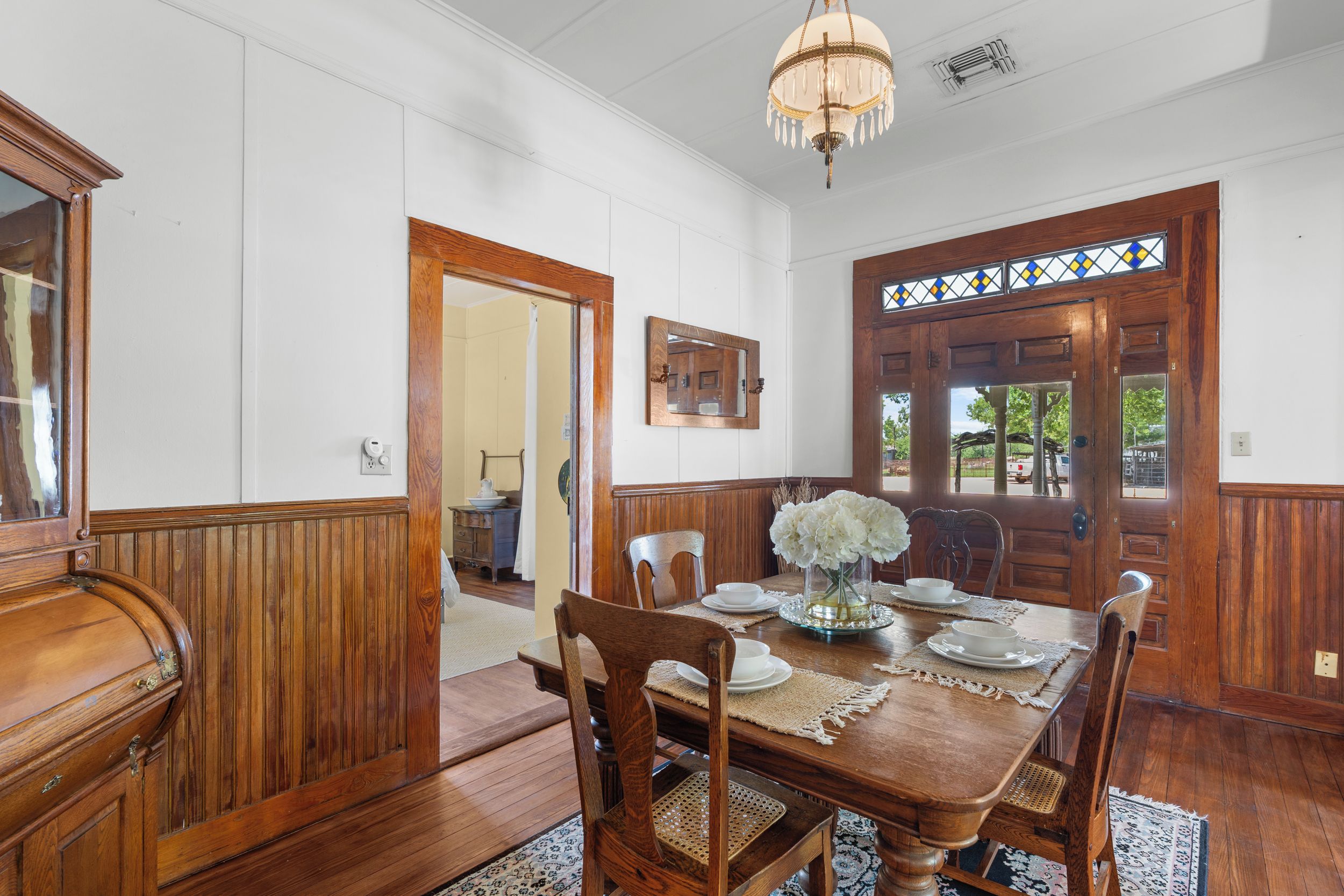 A dining room with a wooden table set for six, surrounded by wooden chairs, a chandelier, and a stained glass window.