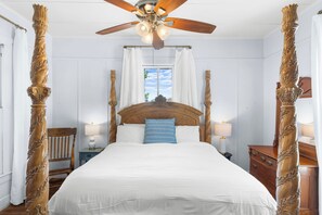 Un dormitorio con una cama grande, columnas de madera decorativas, un ventilador de techo y una ventana con cortinas.