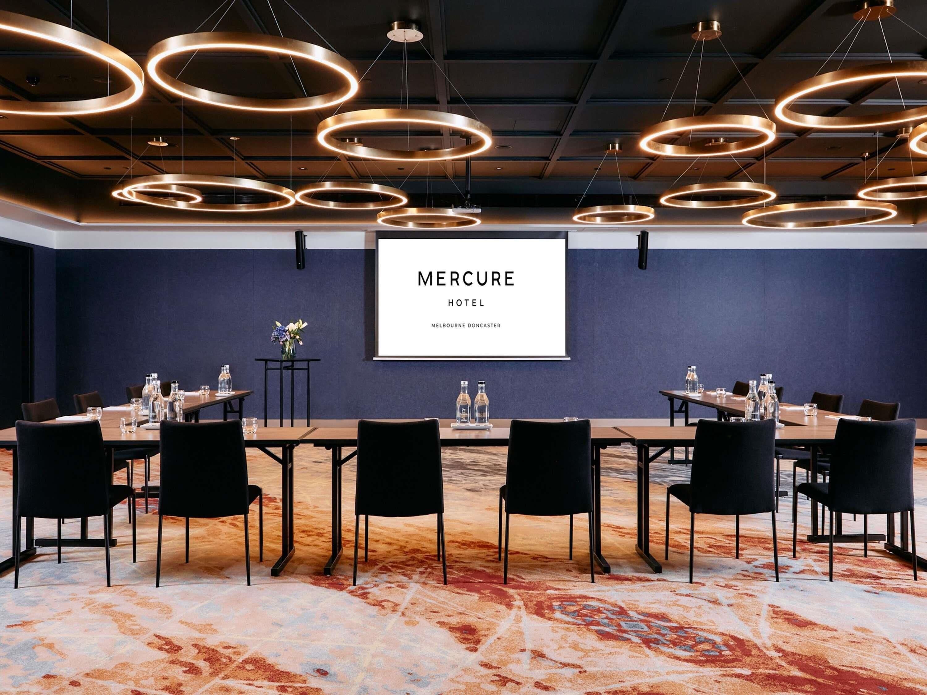 A conference room with a large screen displaying 'Mercure Hotel Melbourne Doncaster,' a long table with chairs, and a patterned carpet.