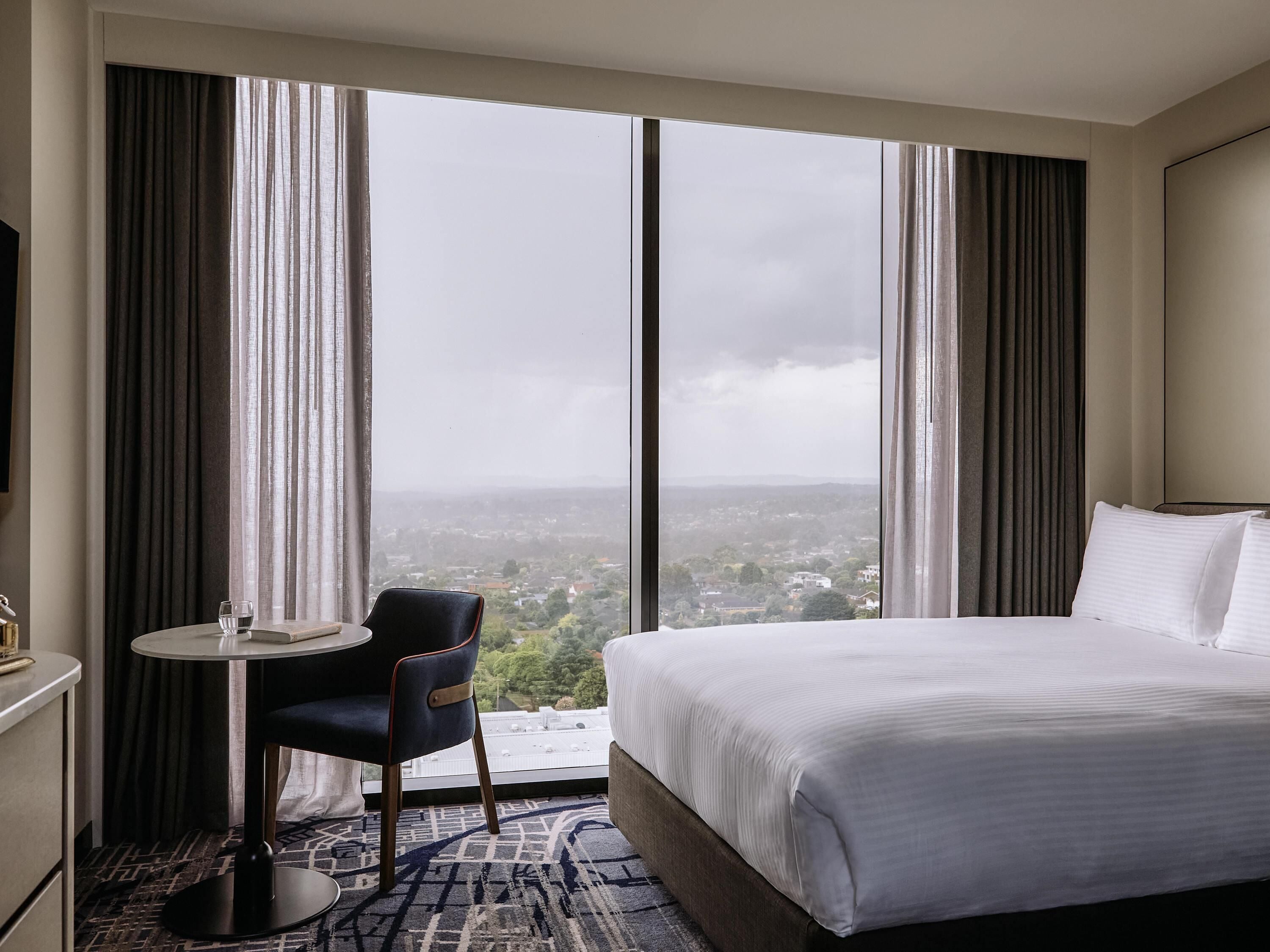 A hotel room with a large bed, a small table with a chair, and a view of a cityscape through large windows.